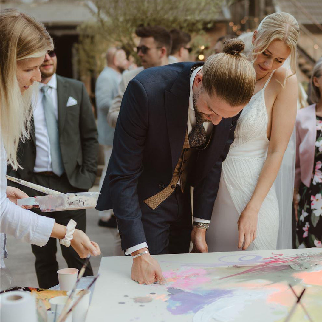 Braut, Bräutigam und Hochzeitsgäste gestalten gemeinsam ein abstraktes Kunstwerk bei einem Gemeinschaftsgemälde.